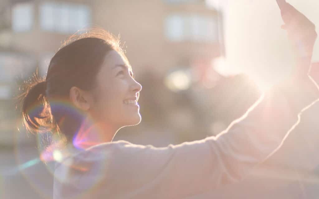 Fotografia de mulher de perfil tirando selfie, sol ao fundo