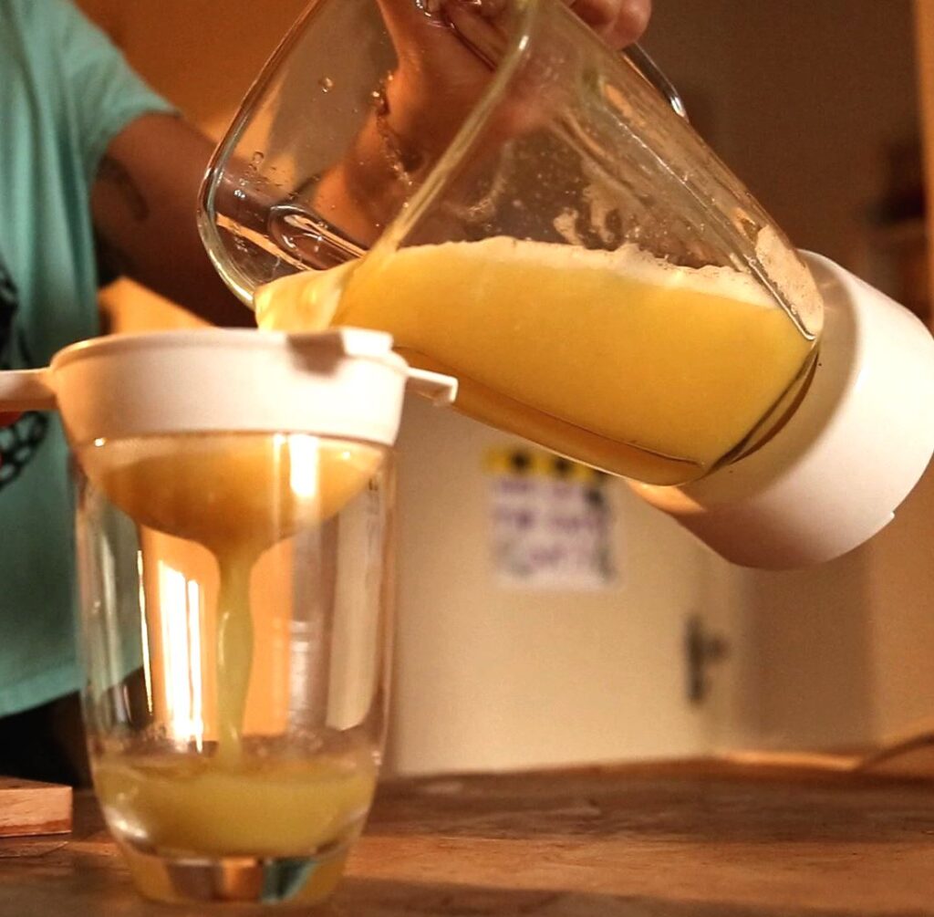 Fotografia de suco amarelo sendo coado do liquidificador para um copo de vidro