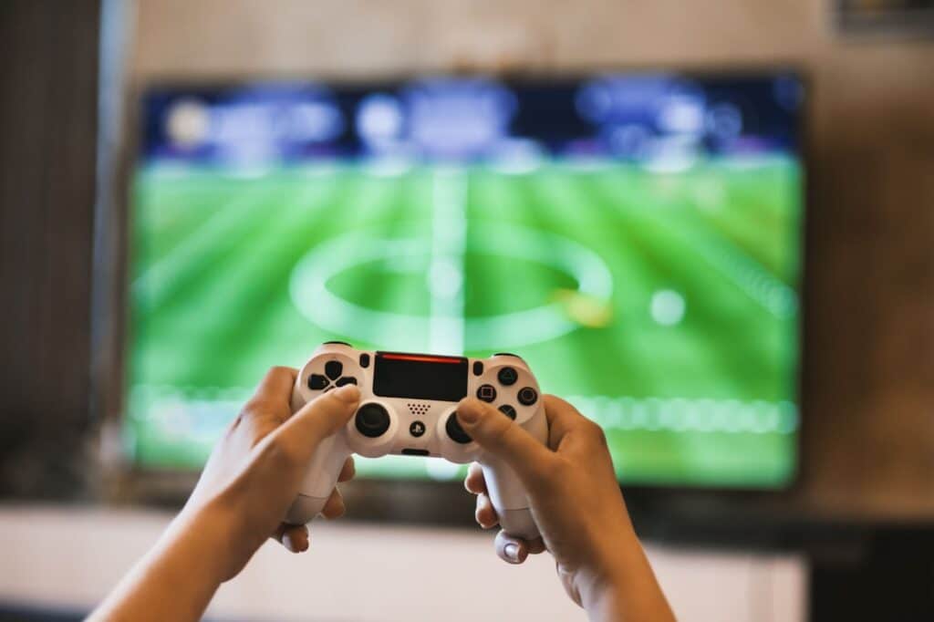 Fotografia de mãos segurando controle remoto. Ao fundo televisão com campo de futebol